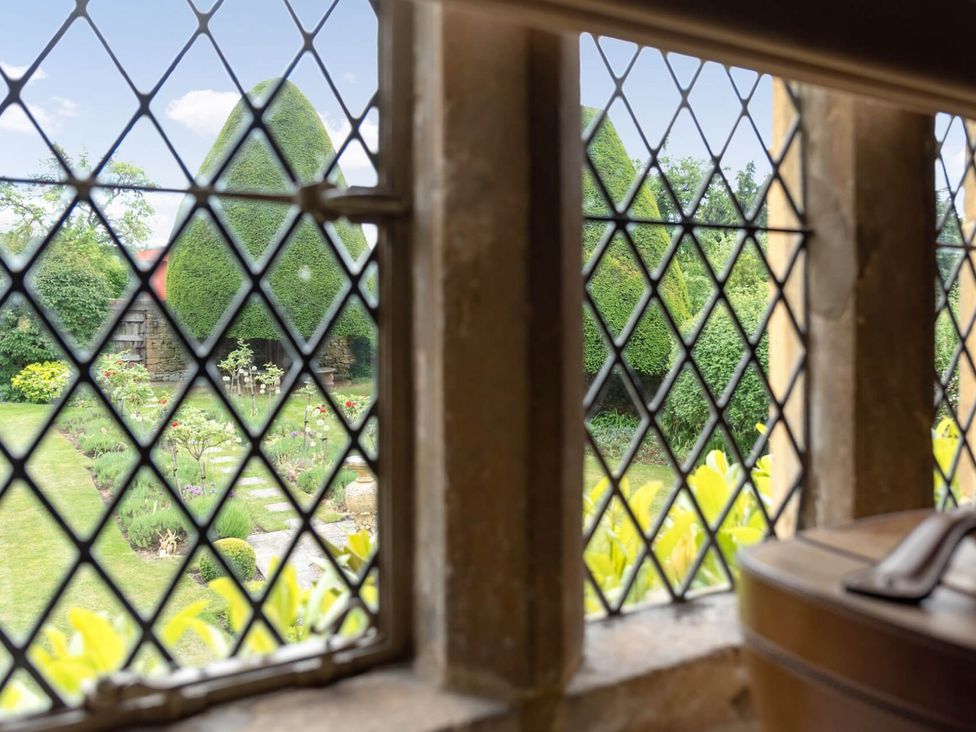 A garden with trimmed topiary trees and flower beds viewed through diamond-paned windows at Little Gables in Broadway
