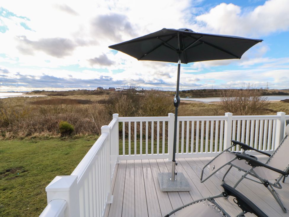 A deck with two lounge chairs and a large umbrella overlooking grassy land and water at Wansbeck View 2 in Ashington