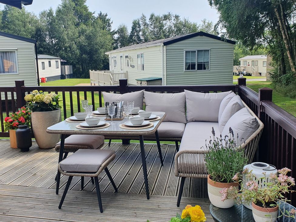 An outdoor dining area with a table set for six and potted plants on a wooden deck with mobile homes in the background at The Treeline in Swarland near Felton