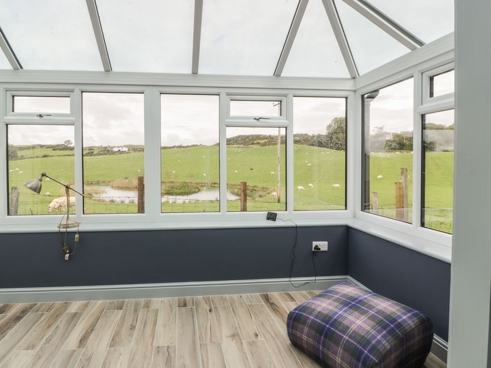 A sunroom with large windows overlooking green fields and a plaid cushioned stool on a wooden floor at Hardhills Cottage in Castle Douglas
