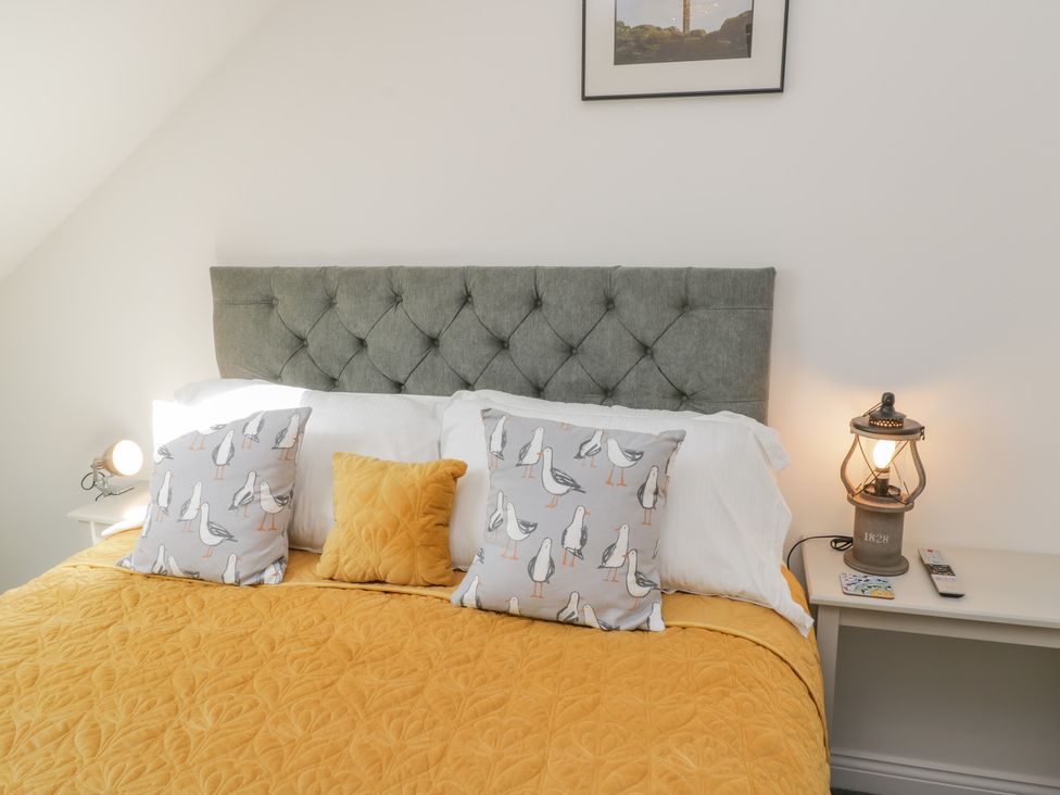 A bed with a grey tufted headboard yellow quilt and pillows with bird patterns and bedside tables with lamps at Foxton Penthouse in Whitby