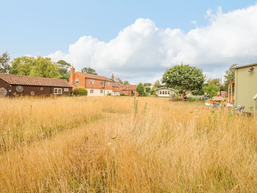 A grassy field with a red brick house and wooden buildings with trees in the background at Willow Cottage in Hickling near Catfield