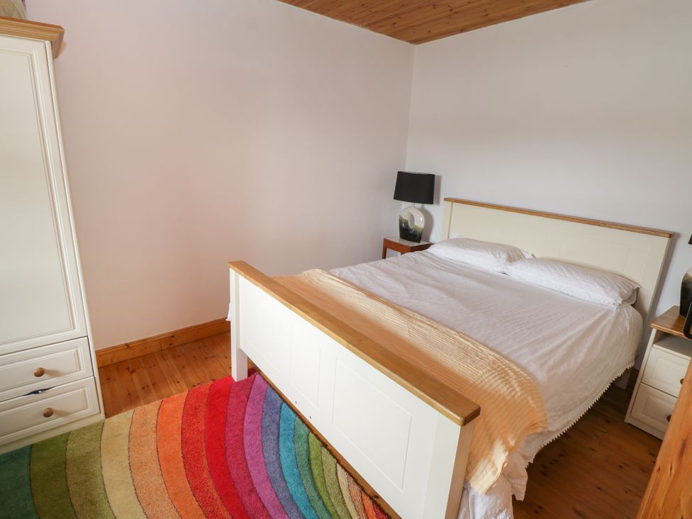A bedroom with a white bed frame white bedding two bedside tables with lamps a white wardrobe and a colorful striped rug on a wooden floor at Tom's Cottage in Rosmoney near Westport County Mayo