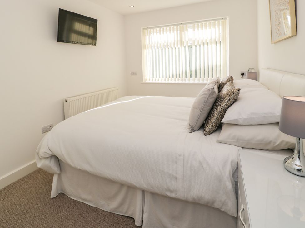A bedroom with a double bed pillows bedside table with lamp wall mounted tv and window with vertical blinds at Top Of The Lane Luxury Holiday Apartment in Benllech