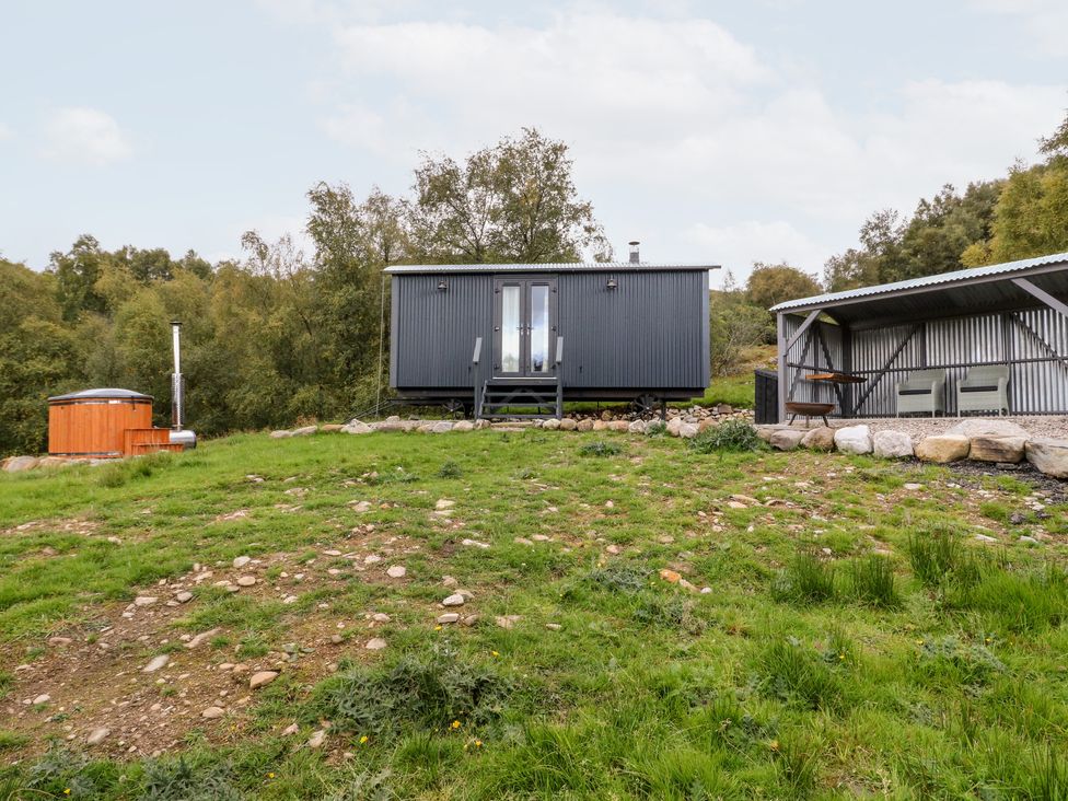 A small black cabin on wheels with stairs leading to the door a wooden hot tub and a covered seating area with two chairs in a grassy rural area at Coraldie Laggan near Newtonmore