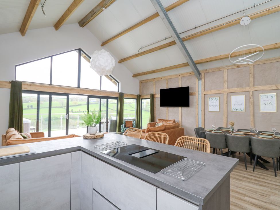 An open plan kitchen and living area with stools at the kitchen island brown sofas a dining table and large windows showing a green landscape at Barcud Coch - Red Kite in Garthmyl
