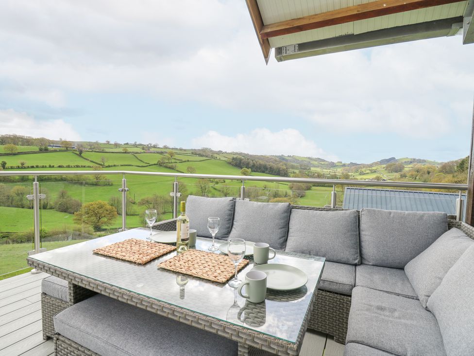 An outdoor seating area with a glass table set with cups plates and wine glasses overlooking green fields at Barcud Coch - Red Kite in Garthmyl