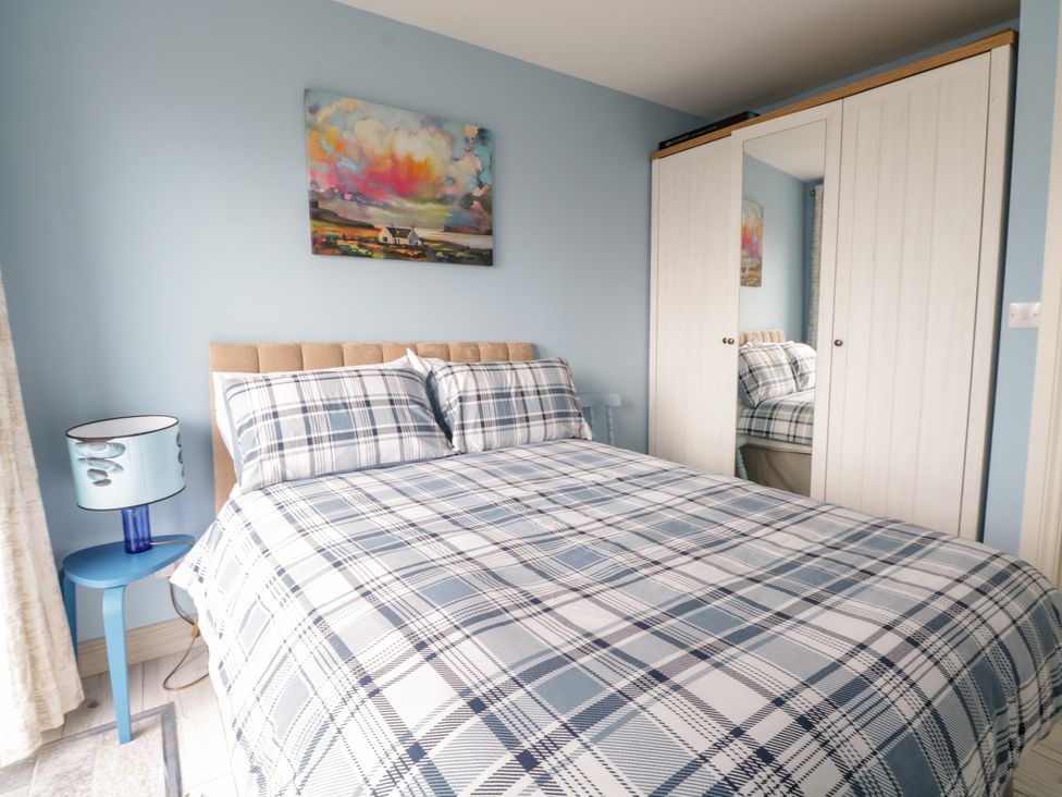 A bedroom with a bed in plaid bedding a blue side table and lamp a wardrobe with mirror and a painting on the wall at 38 Carrowhubbock Holiday Village in Enniscrone County Sligo