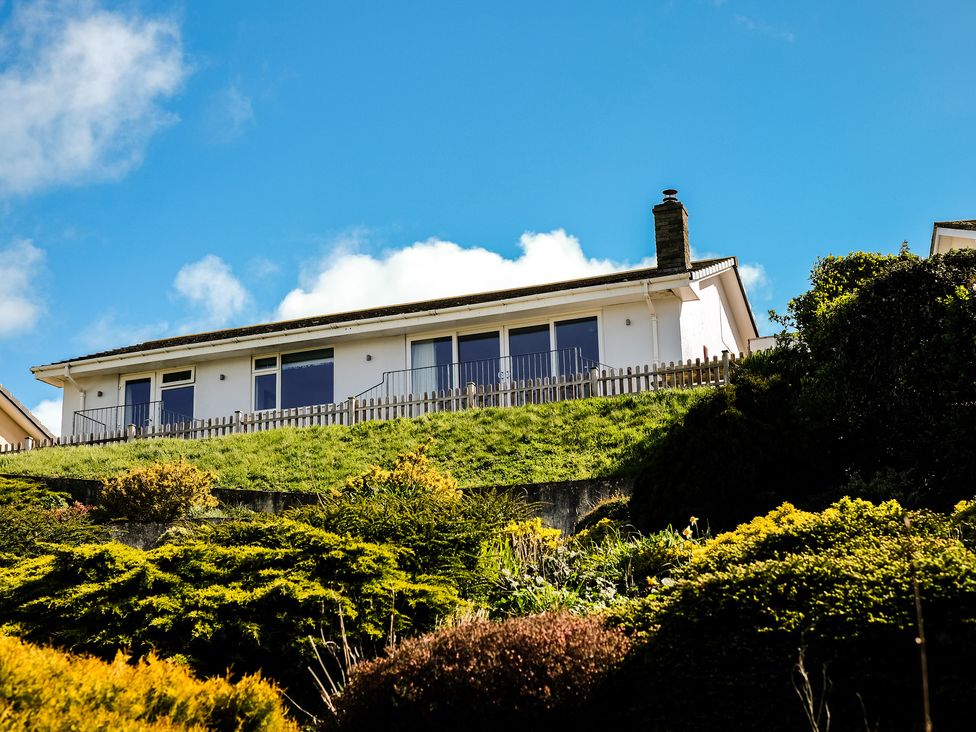 A white house with large glass windows on a grassy hill surrounded by bushes at Harbour Lights Port Isaac