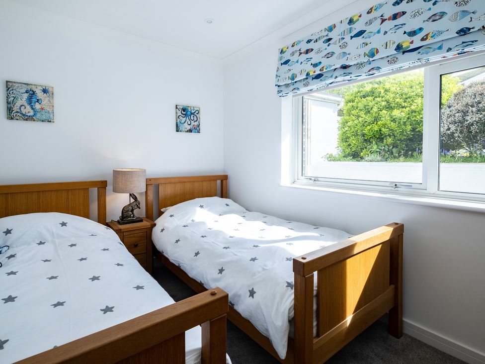 A bedroom with two single wooden beds white star patterned bedding a bedside table with a lamp and window with fish patterned blind at Harbour Lights Port Isaac