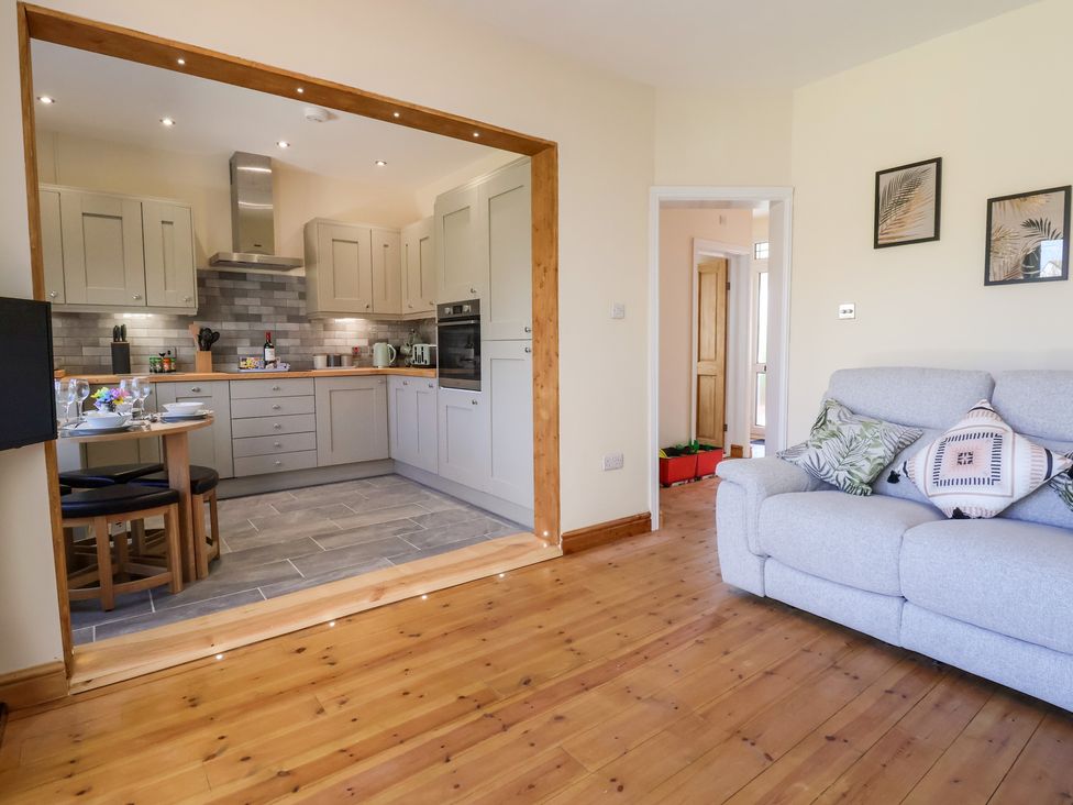A living room with a gray sofa and pillows next to an open kitchen with cabinets and a dining table at 3 Rhyd Drive in Rhos-On-Sea