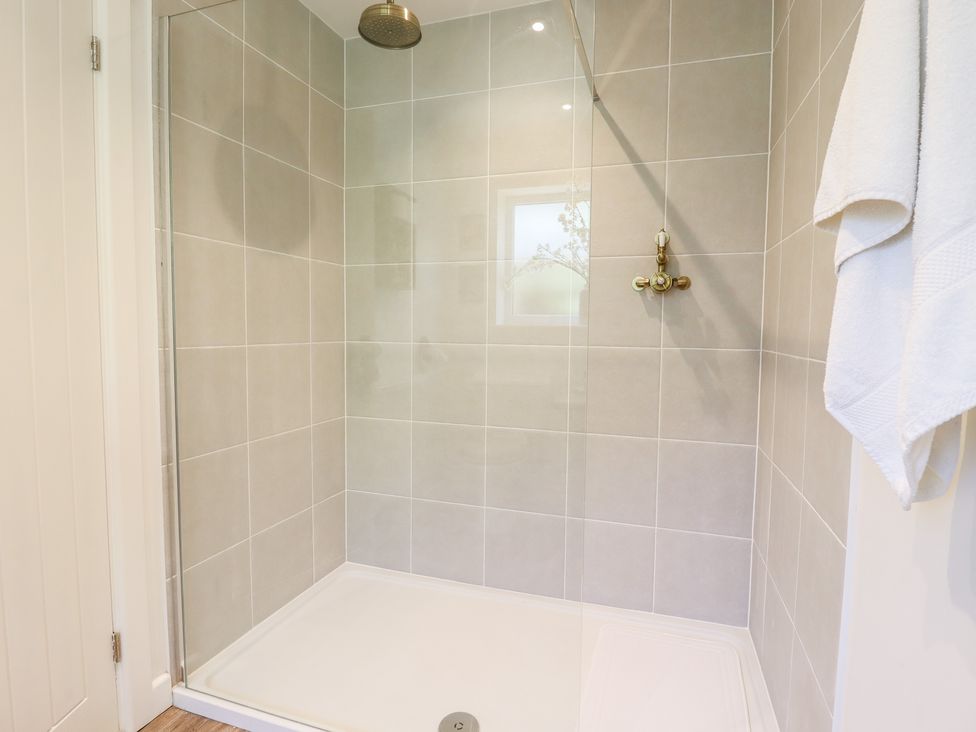 A tiled shower area with a glass door a brass shower head and controls a small window and a white towel hanging at Pantiles in Eyke near Woodbridge