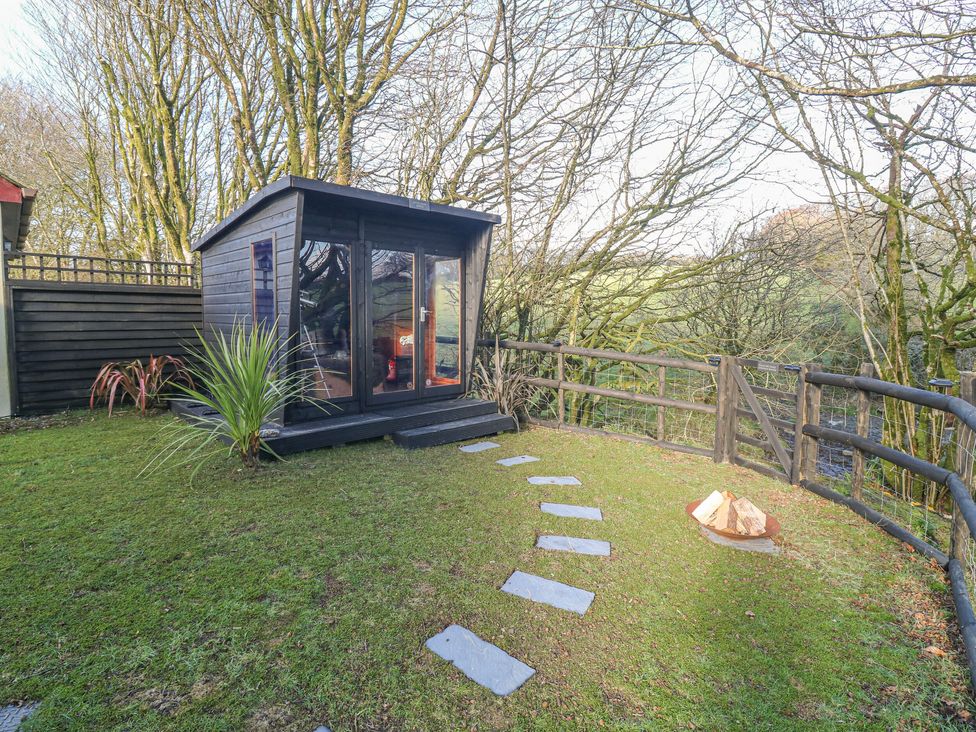 A small black wooden cabin with glass doors in a fenced garden with a stone pathway and firewood pile at Inny Brook Cottage in Davidstow near Camelford