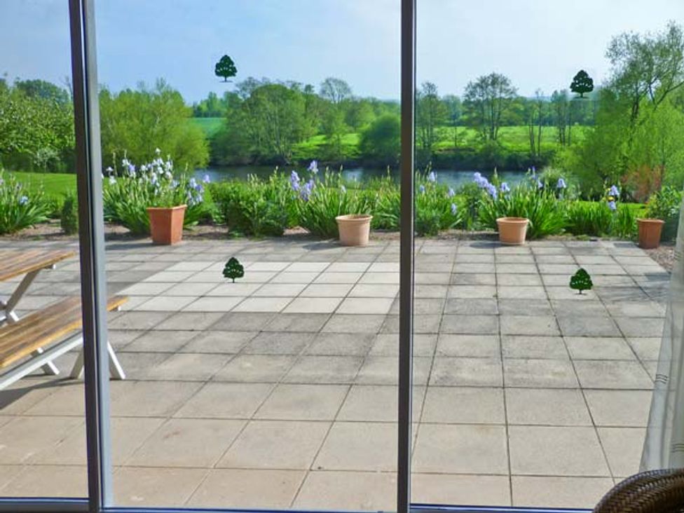 A patio with flower pots and a picnic bench overlooking a garden and river at Herons in Byford