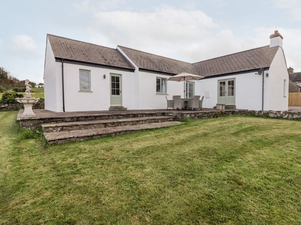 A white house with stone steps leading to a patio with table chairs and umbrella and a green lawn at Fountain Folly in Moylgrove near St Dogmaels