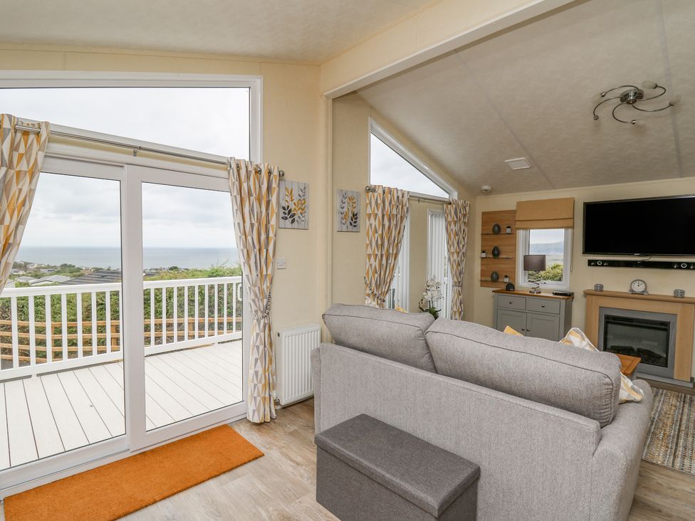 A living room with a gray sofa a wall-mounted tv and a fireplace next to sliding glass doors leading to a deck with a sea view at Platinum Lodge in Borth