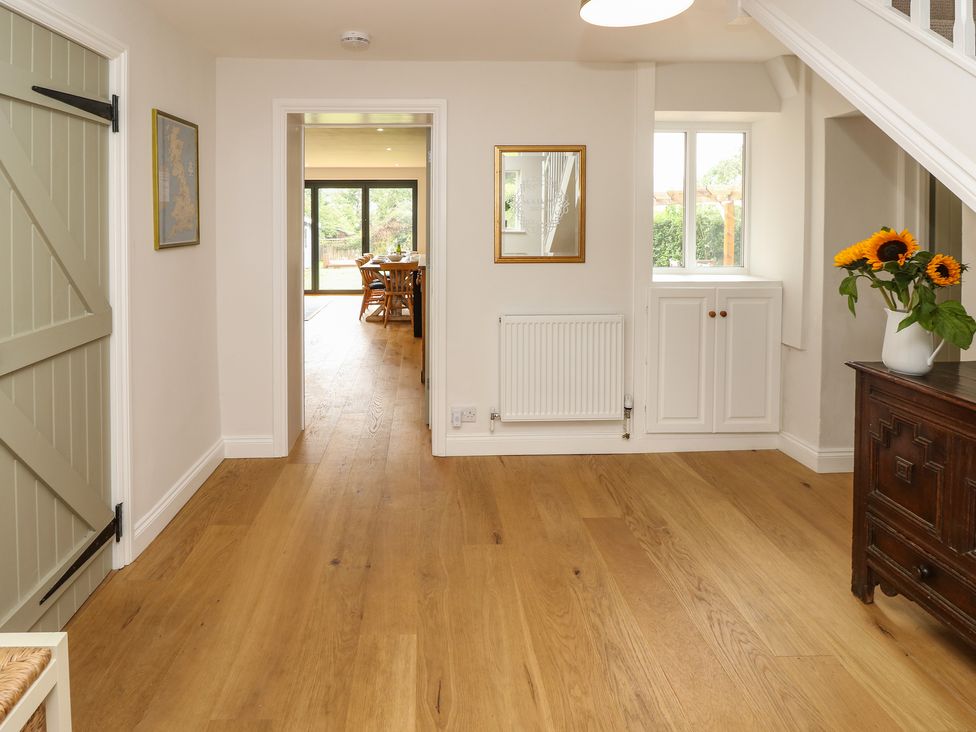 A hallway with wooden floors a green door a framed map a radiator a mirror a window a white cabinet a dark wooden sideboard with sunflowers and a view into a dining area at Frosthill Cottage in Carisbrooke