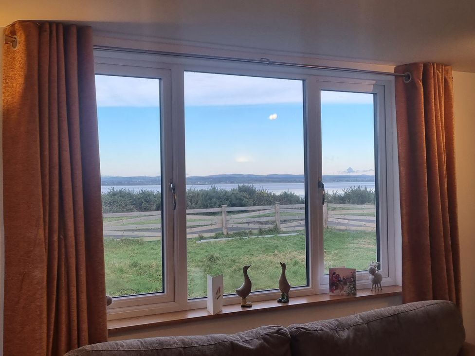 A window with brown curtains looking out to grass a wooden fence and water at Cherry Croft in Bowness-On-Solway