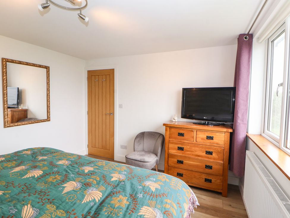 A bedroom with a bed covered by a patterned blanket a wooden dresser with a TV and a chair next to a wooden door at Cherry Croft in Bowness-On-Solway
