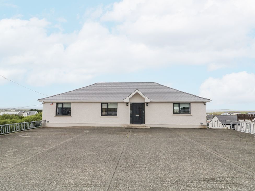 A single-story house with a dark front door and three windows facing a large concrete driveway at Cottage Catriona in Derrybeg County Donegal