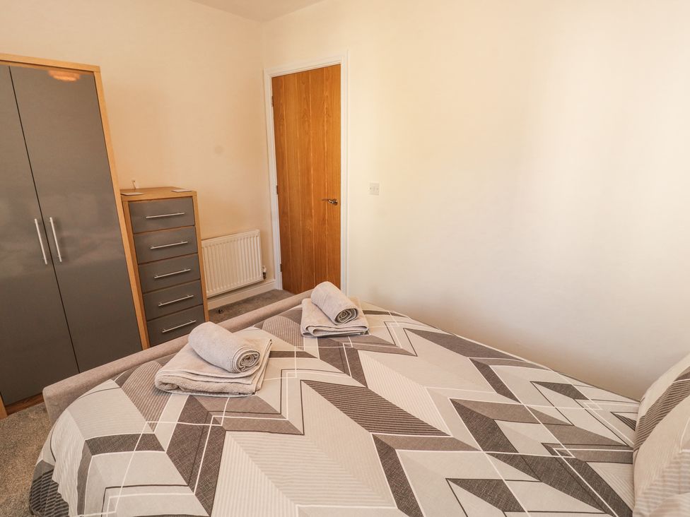 A bedroom with a bed covered in a geometric patterned duvet two rolled towels on the bed a wardrobe and a chest of drawers near a wooden door at Squirrel Cottage in Penruddock