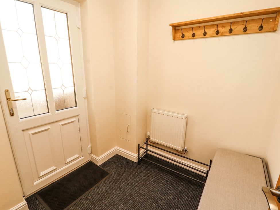 An entryway with a white door a black floor mat a radiator a metal shoe rack a wooden wall-mounted coat rack and a bench at Squirrel Cottage in Penruddock