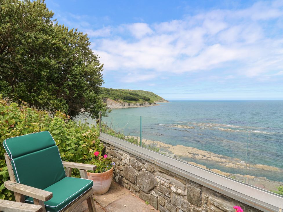 A wooden chair with green cushions on a patio next to a flowerpot and glass railing overlooking the sea and cliffs at Pen y Graig in Aberporth