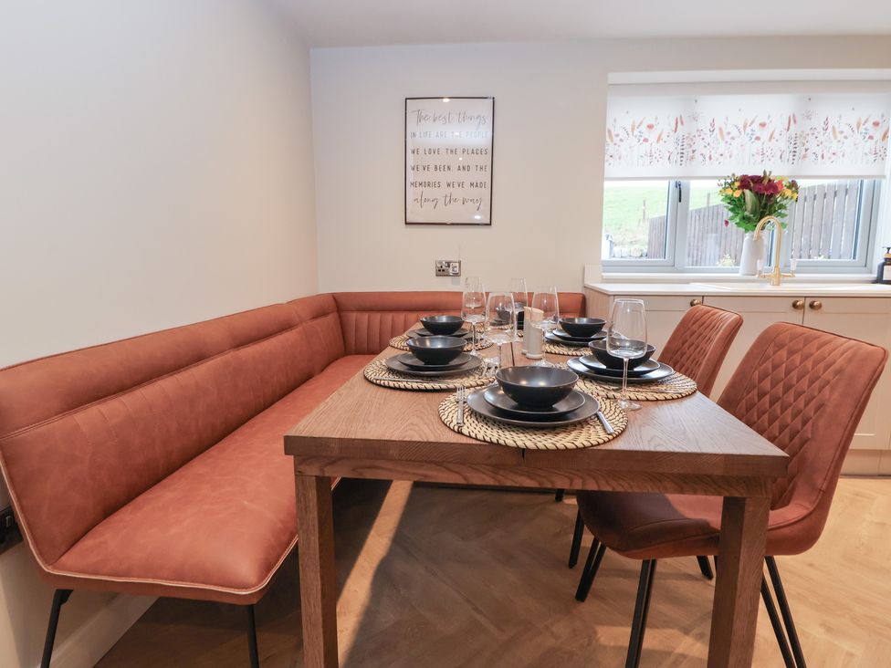 A dining area with a wooden table set with black dishes and glassware plus brown cushioned bench and chairs near a window with floral curtains at Beudy Pwll Pridd in Llangollen