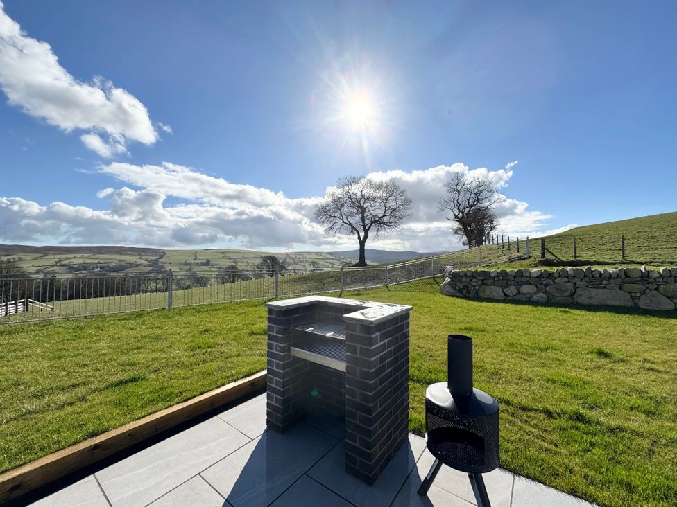 An outdoor patio with a brick grill and metal fire pit with grassy fields and hills in the background at Beudy Pwll Pridd in Llangollen