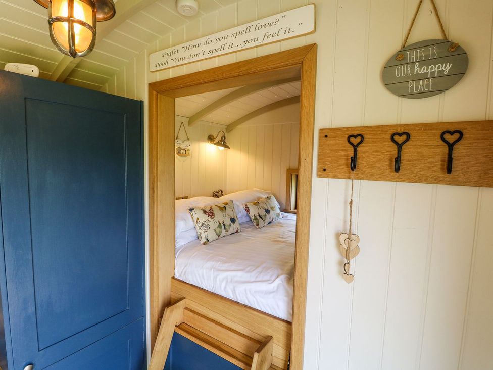 A bedroom with a bed with white sheets and decorative pillows viewed through a wooden doorframe with a blue door and wall hooks on a white paneled wall at Lillies Shepherds Hut at Shepherds Lodge Bottesford near Redmile and Vale of Belvoir