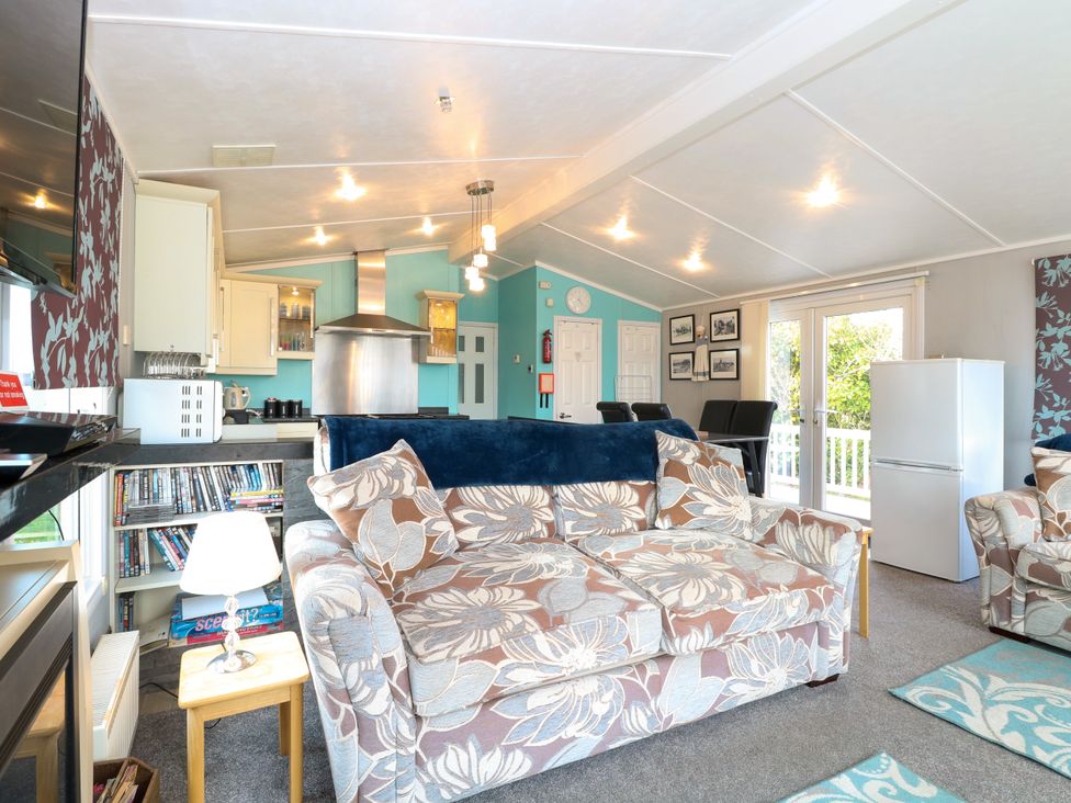 A living room with floral sofas a small table with a lamp a bookcase and a kitchen area at 3 Bure Close in Burgh Castle