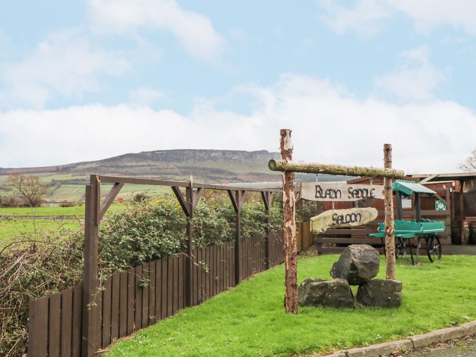 A wooden sign reading Blazin Saddle Saloon on grass near a wooden fence and green cart with hills in the background at Bear Claw Lodge in Castlerock