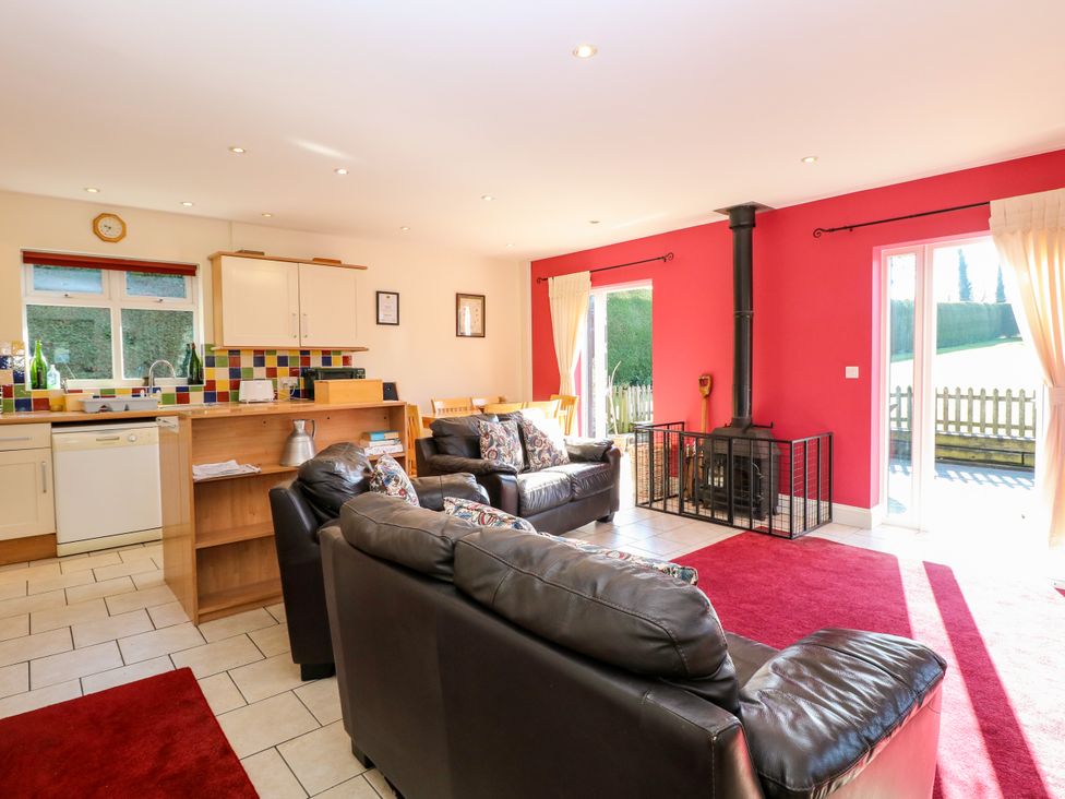 An open living area with black leather sofas a wood stove with protective screen red walls and an adjacent kitchen with tiled backsplash and white cabinets at Swallow Barn in Holton near Halesworth