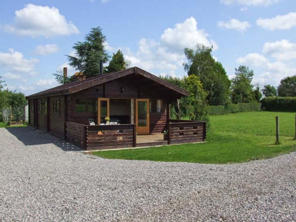 A wooden cabin with a porch and gravel driveway surrounded by grass and trees at Cropvale Lodge in Cropthorne near Pershore