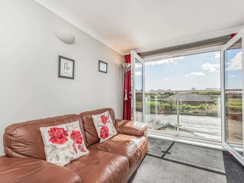 A room with a brown leather sofa and floral cushions next to glass doors overlooking a balcony and outdoor view at 16 The Boathouse in Rye