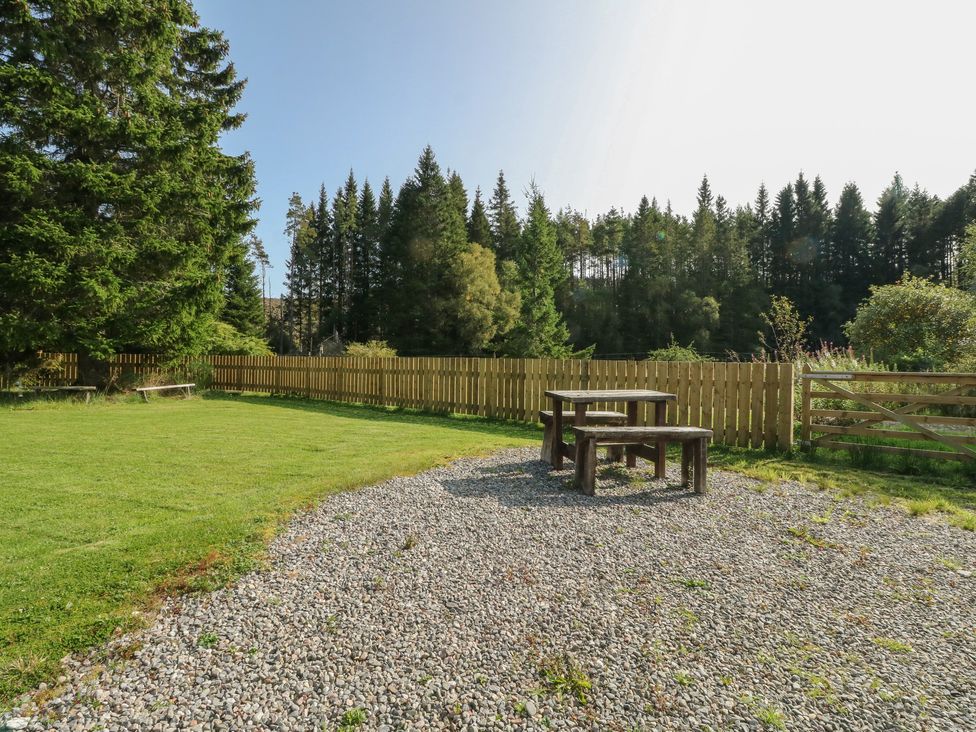 An outdoor area with a wooden table and benches on gravel next to a grassy lawn and fenced trees at Feagour in Laggan
