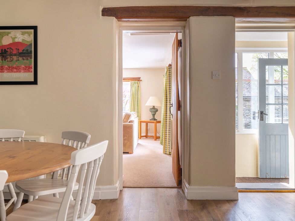 A dining area with a wooden table and white chairs and a doorway leading to a living room with a lamp on a side table and a door opening to the outside at Bassicks Hawkshead
