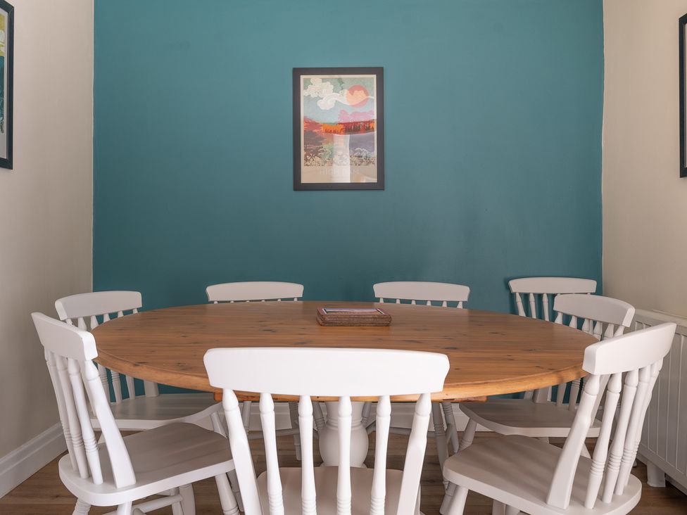 A dining room with a round wooden table and six white chairs with a framed picture on a teal wall at Bassicks in Hawkshead
