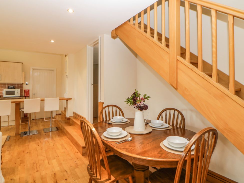 A dining area with a wooden table set with dishes and cutlery under a wooden staircase near a kitchen with bar stools at Meader Barn Peter Tavy near Tavistock