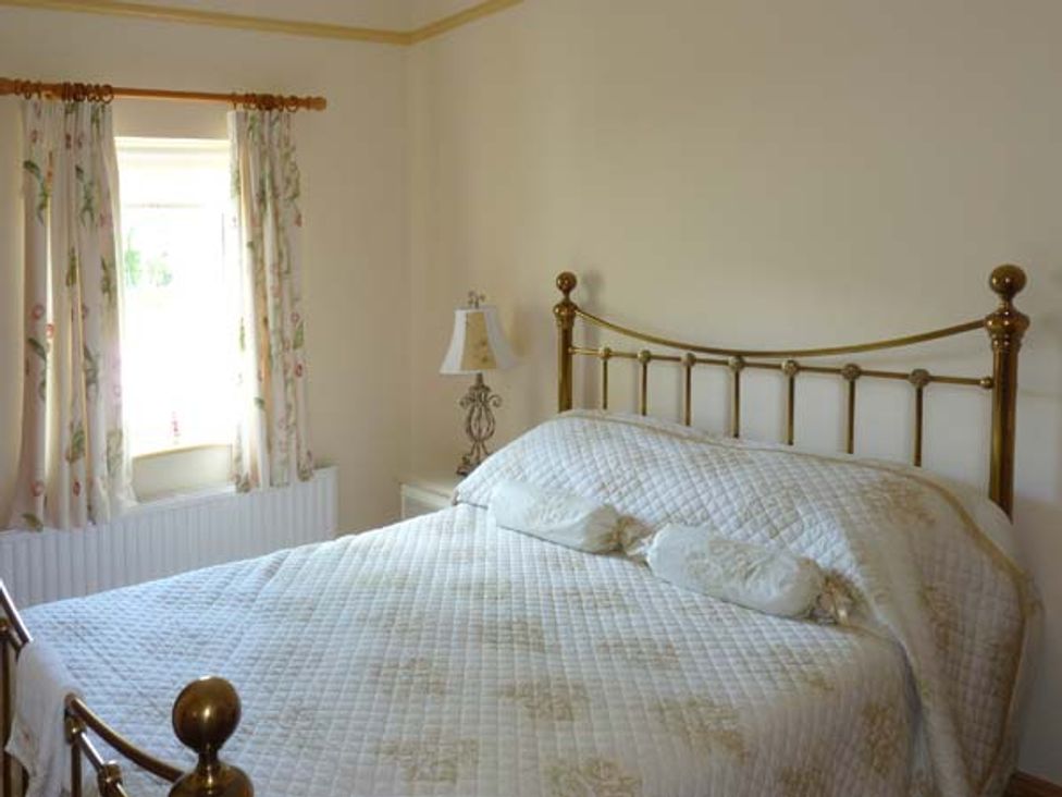 A bedroom with a brass bed frame, a quilted bedspread, a bedside table with a lamp, and a window with floral curtains at Annies Cottage in Doonamona near Belcarra County Mayo