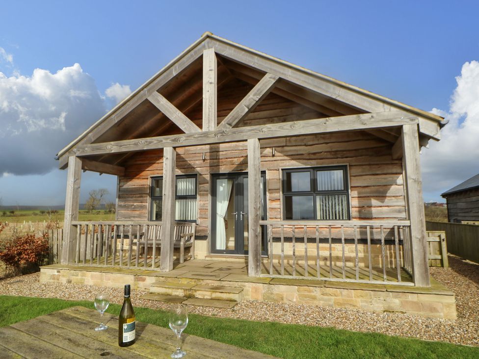A wooden cabin with a porch bench and table with a wine bottle and glasses on the grass at Coquet in Longframlington near Alnwick