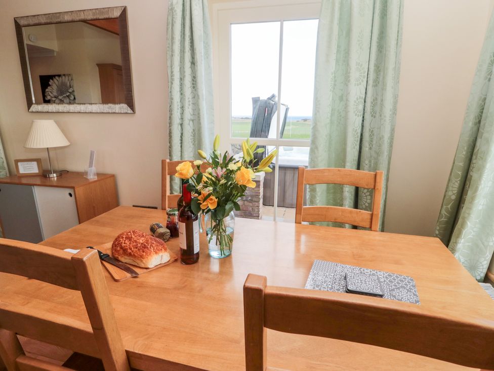 A dining room with a wooden table and chairs a bouquet of flowers bread and wine on the table and a window with green curtains at Bank Top Cottage in Embleton