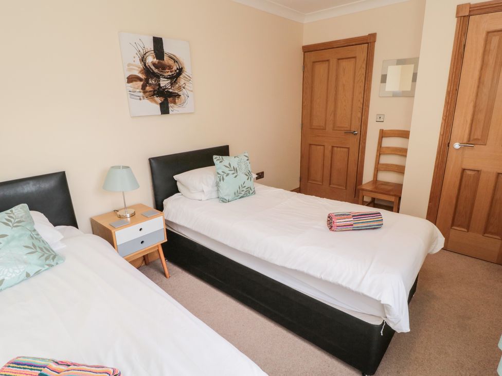 A bedroom with two single beds a nightstand with a lamp two wooden doors a wooden chair and abstract wall art at Bank Top Cottage in Embleton