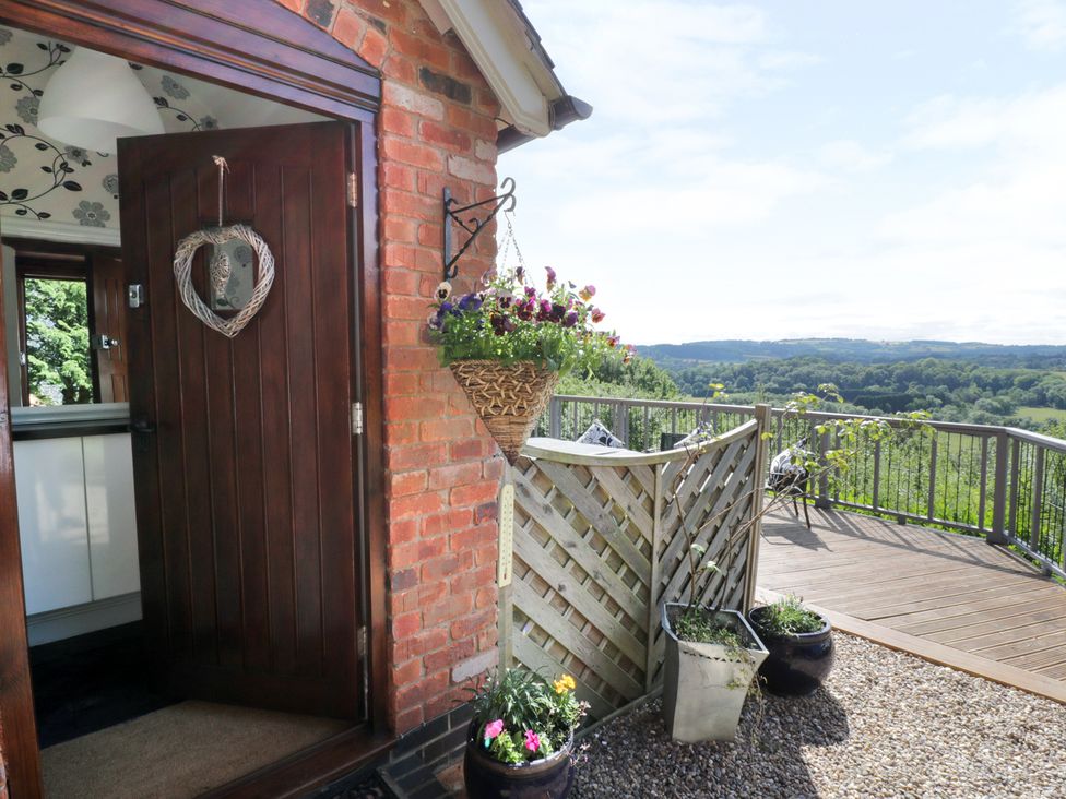 An open wooden door on a brick wall leading to an interior with floral wallpaper a hanging flower basket and potted plants on a gravel floor and a wooden deck with a metal railing overlooking a green landscape at Hillview Cottage in Knightwick near Worcester