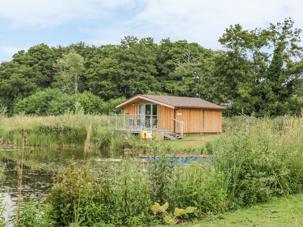 A wooden cabin by a pond with surrounding greenery and trees at Lakeside Lodge in East Harling near Thetford