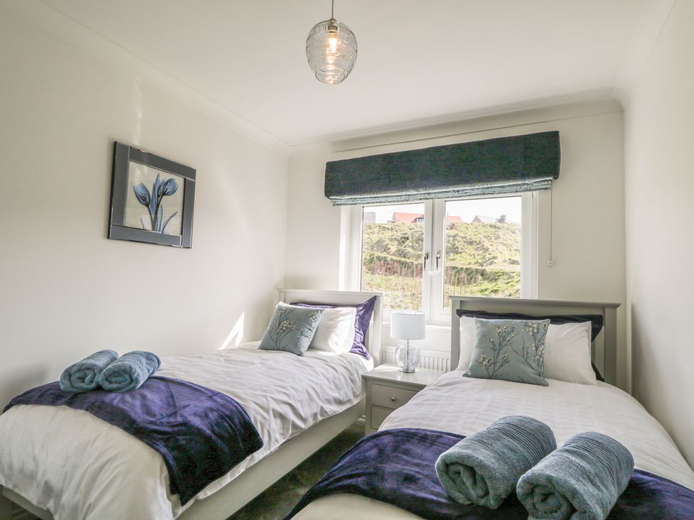 A bedroom with two single beds, a nightstand with a lamp between them, a window with a dark valance, and a framed flower artwork on the wall at 24 Waters Edge in Banff