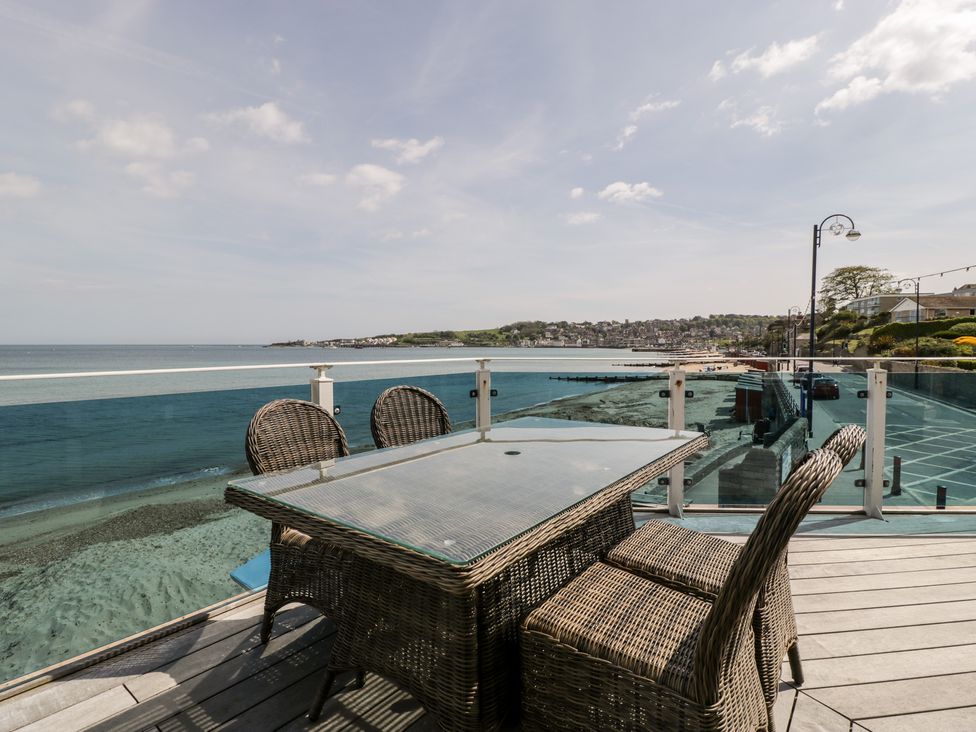 A balcony with a glass table and wicker chairs overlooking the sea and a coastal town at 1 Ocean Bay in Swanage