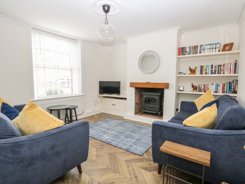 A living room with two blue sofas with yellow pillows a fireplace a TV on a stand bookshelves with books and decorations and a window with blinds at Springfield in Hempton near Fakenham