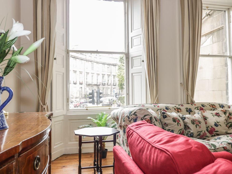 A sitting area with floral and red sofas next to a window with beige curtains and a wooden side table at 3 Lynedoch Place in Edinburgh
