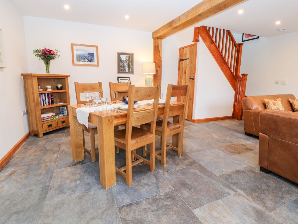 A dining area with a wooden table and chairs a wooden cabinet with books and flowers a staircase and a brown leather sofa at The Old Cart House in Nantmawr near Trefonen